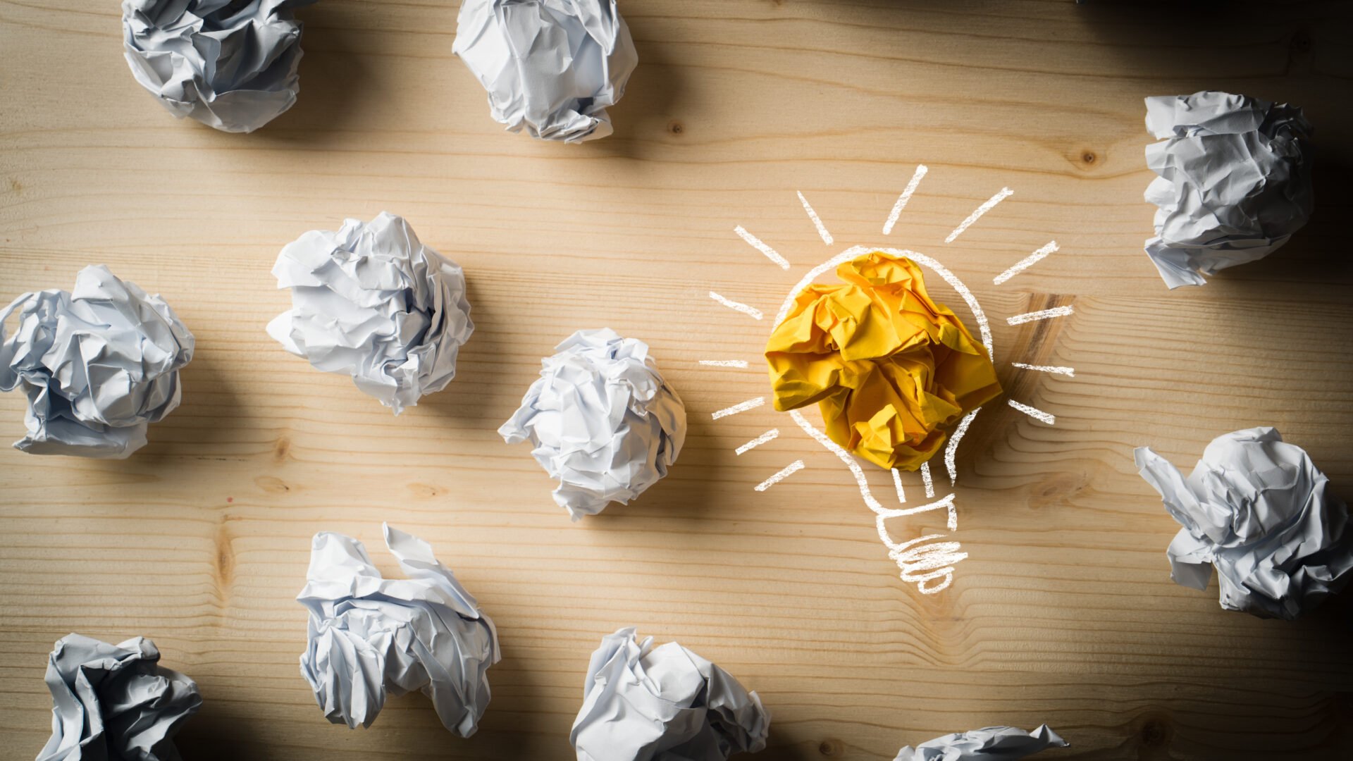 Weiße, zusammengefaltete Papierkugeln, gelbe Papierkugel mit Umriss einer Glühbirne